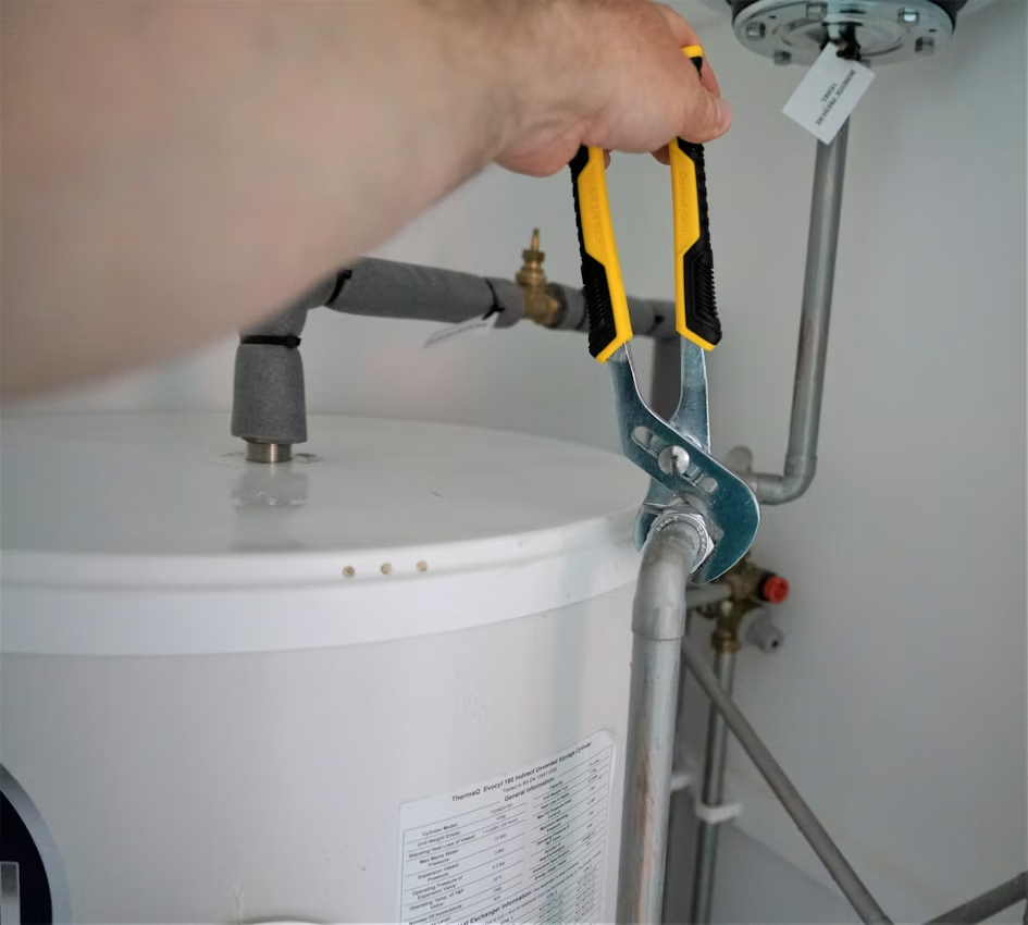 A close-up of a person using yellow-handled pliers to tighten or loosen a pipe fitting on a white residential water heater unit.  